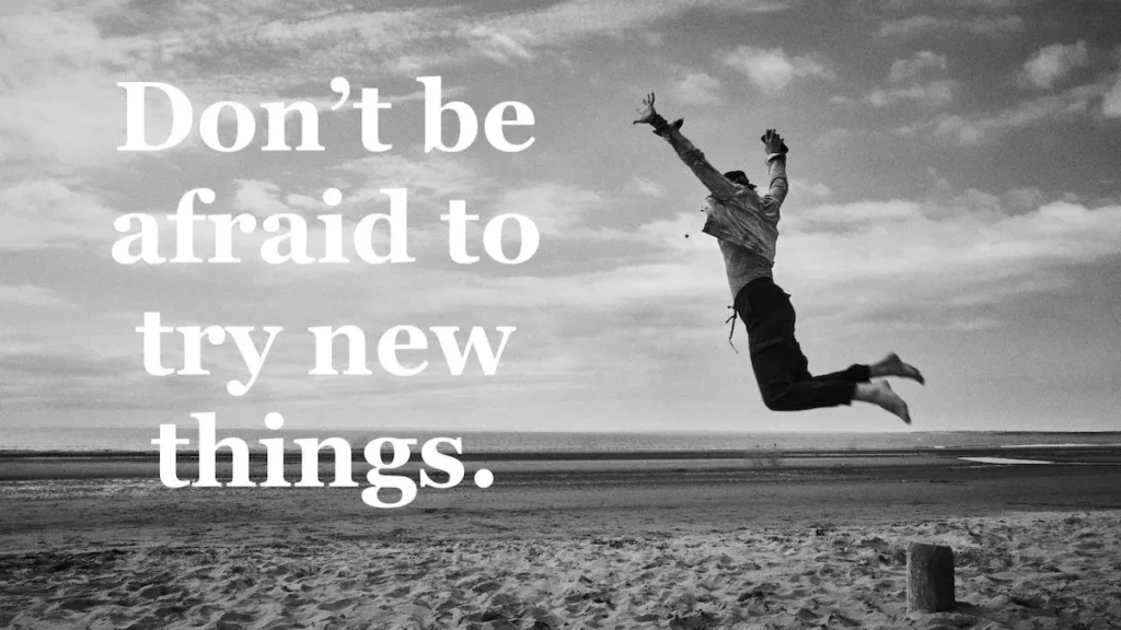 Black and white picture of a person jumping into the air on a beach. Quote: Don't be afraid to try new things.