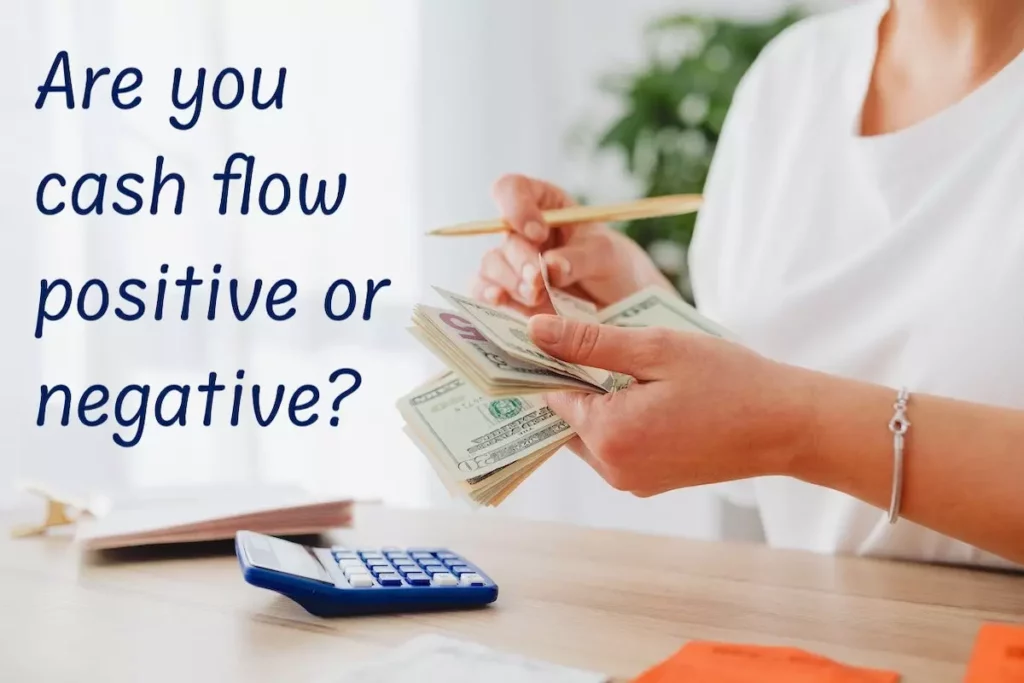 Image of a person with a calculator, counting dollar bills with the text overlay: Are you cash flow positive or negative?
